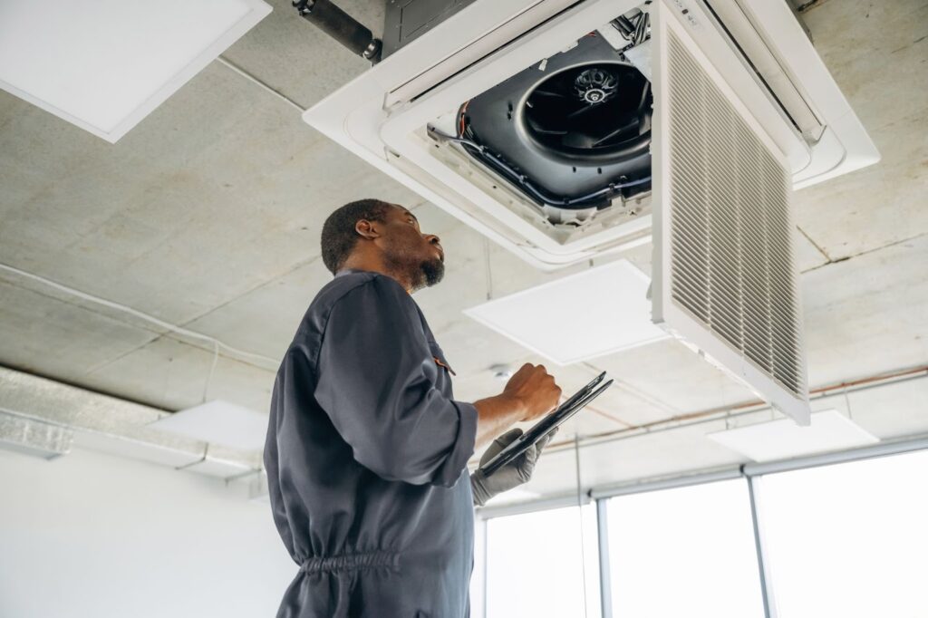 Technician in a gray uniform inspecting an office air conditioning unit and recording findings on a clipboard